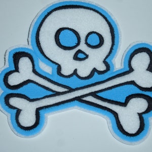Könnte beinhalten: Gestickter Aufnäher mit einem weißen Totenkopf und gekreuzten Knochen, umrandet in Schwarz. Das Design hat einen hellblauen Hintergrund und einen weißen Rand. Ein klassisches Piraten-Themen-Emblem.