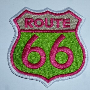 Könnte beinhalten: Gestickter Route 66-Aufnäher in Schildform. Der Aufnäher hat einen grünen Hintergrund mit der Zahl "66" in Pink und dem Wort "ROUTE" in Pink darüber. Der Rand ist weiß und pink.