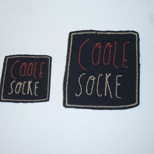 Könnte beinhalten: Zwei schwarze quadratische Aufnäher mit den Worten "COOLE SOCKE" in roter und cremefarbener Stickerei. Die Aufnäher haben einen cremefarbenen Rand und befinden sich auf weißem Hintergrund.