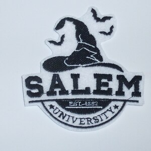 Könnte beinhalten: Ein schwarz-weißer, gestickter Aufnäher mit einem Hexenhut und Fledermäusen über dem Text "SALEM UNIVERSITY EST. 1692". Der Aufnäher hat ein kreisförmiges Design mit einem Stern auf jeder Seite des Wortes "UNIVERSITY".