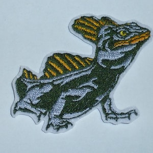 Könnte beinhalten: Gestickter Aufnäher mit einer stilisierten Eidechse mit einer markanten Rückenflosse. Der Aufnäher hat einen dunkelgrünen Körper mit silbernen und goldenen Akzenten und ein gelbes Auge. Der Aufnäher befindet sich auf weißem Hintergrund.