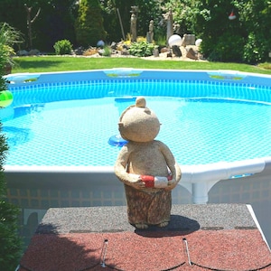 May include: A beige garden statue of a person holding a red and white life preserver stands in front of a blue swimming pool. The pool is surrounded by a green lawn and trees. A green ball floats in the pool.
