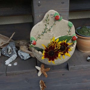 Könnte beinhalten: Ein Keramik-Wandtopf mit beigefarbenem Hintergrund und floralem Design. Der Topf hat einen erhöhten Rand und einen Ausschnitt in Form einer Blume. Der Topf ist mit Marienkäfern und Sonnenblumen verziert.
