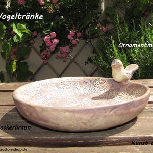 Könnte beinhalten: Ockerbraunes Vogelbad aus Keramik mit dekorativem Blumenmuster. Eine kleine Vogelfigur sitzt am Rand. Das Vogelbad steht auf einer Holzoberfläche, im Hintergrund Grün und Blumen.