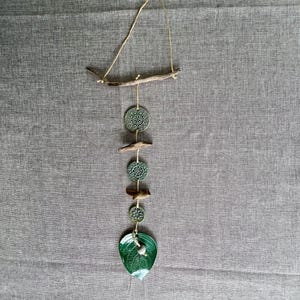 Puede incluir: Un colgante de pared artesanal con una rama de madera natural y una cuerda de yute para colgar. El diseño incluye elementos de cerámica verde: piezas circulares con un patrón floral y una forma de hoja. La decoración de la pared es una pieza inspirada en la naturaleza.
