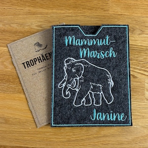 Könnte beinhalten: Eine dunkelgraue Filzhülle mit einer weißen Mammut-Stickerei, dem türkisfarbenen Schriftzug "Mammut-Marsch" und "Janine" sowie einem türkisfarbenen Rand. Daneben liegt ein beigefarbenes Heft mit dem Text "TROPHÄEN" und einer kleinen Stiefelgrafik.