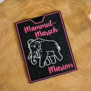 Könnte beinhalten: Schwarzer Filz-Patch mit rosa Nähten. Der Patch zeigt ein weiß gesticktes Bild eines Mammuts mit dem Text "Mammut-Marsch Marion" in rosa.