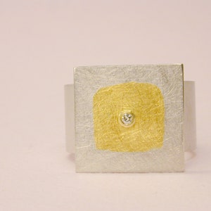 Könnte beinhalten: Ein silberner Ring mit quadratischem Design. Die Mitte des Rings ist ein goldenes Quadrat mit einem kleinen Diamanten in der Mitte.