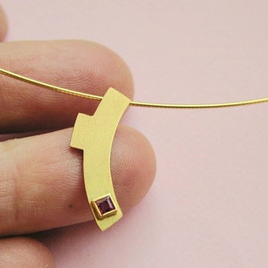 Può includere: Una collana d'oro con un ciondolo geometrico con una piccola pietra rossa quadrata.