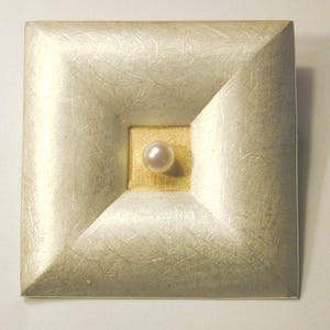 Könnte beinhalten: Quadratische, silberfarbene Brosche mit einem zentralen goldfarbenen Quadrat. In der Mitte ist eine einzelne Perle eingesetzt. Die Brosche hat eine strukturierte Oberfläche und ein geometrisches Design, geeignet für formelle Kleidung oder als dekoratives Accessoire.