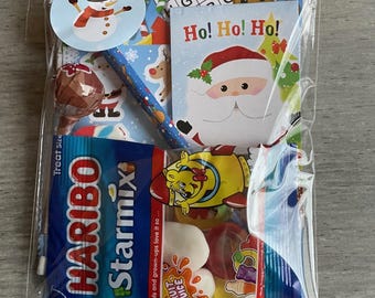 May include: A cellophane bag containing Christmas-themed sweets and small gifts. Includes a snowman sticker, a pencil, a lollipop, a small book with Santa, and a bag of Haribo Starmix sweets. The book says "Ho! Ho! Ho!"