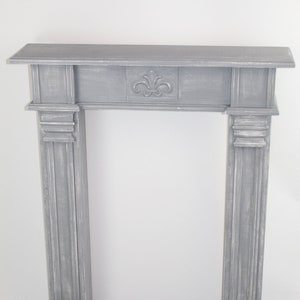 K&ouml;nnte beinhalten: Ein grauer Kaminsims aus Holz mit einem dekorativen geschnitzten Design auf der Oberseite. Der Kaminsims hat eine einfache rechteckige Form und ist aus verwittertem Holz gefertigt.