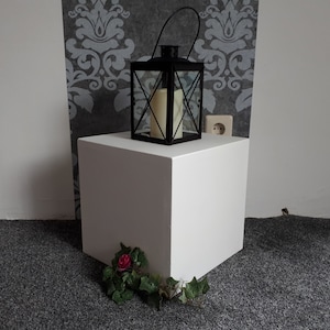 May include: A white cube pedestal with a black lantern and a single white candle on top. A small bouquet of green leaves and a single red rose are in front of the pedestal.