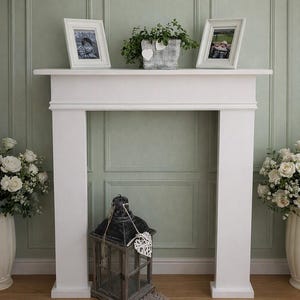 May include: A white wooden fireplace mantel surround featuring a flat top shelf. The mantel is decorated with two white picture frames, a grey planter with green foliage, and a dark grey lantern with a white heart ornament placed on the floor.