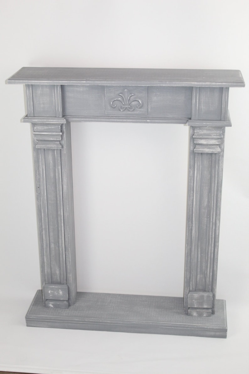 K&ouml;nnte beinhalten: Ein grauer Kaminsims aus Holz mit einem dekorativen geschnitzten Design auf der Oberseite. Der Kaminsims hat eine einfache rechteckige Form und ist aus verwittertem Holz gefertigt.