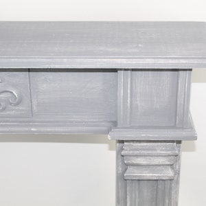 K&ouml;nnte beinhalten: Ein grauer Holzmantel mit einem dekorativen Fleur-de-lis-Design auf der Vorderseite. Der Kaminsims hat ein schlichtes, elegantes Design und w&auml;re eine gro&szlig;artige Erg&auml;nzung f&uuml;r jedes Zuhause.