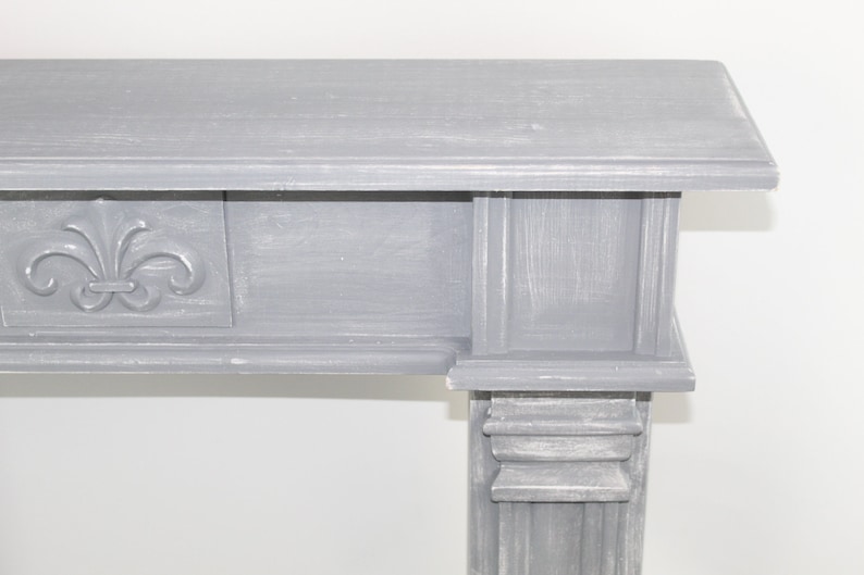 K&ouml;nnte beinhalten: Ein grauer Holzmantel mit einem dekorativen Fleur-de-lis-Design auf der Vorderseite. Der Kaminsims hat ein schlichtes, elegantes Design und w&auml;re eine gro&szlig;artige Erg&auml;nzung f&uuml;r jedes Zuhause.
