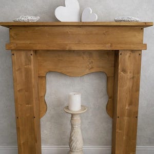 Op de afbeelding: Een bruine houten schouw met een rechthoekige bovenplank en twee verticale zijzuilen. Twee witte hartvormige ornamenten staan op de schouw. Een witte stompkaars staat op een hoge, getextureerde, gebroken witte voetstukstandaard.