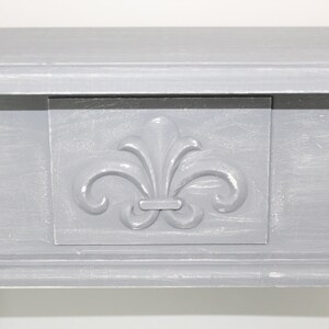 K&ouml;nnte beinhalten: Ein graues Holzregal mit einem dekorativen Fleur-de-lis-Design auf der Vorderseite. Das Regal hat ein schlichtes, elegantes Design.