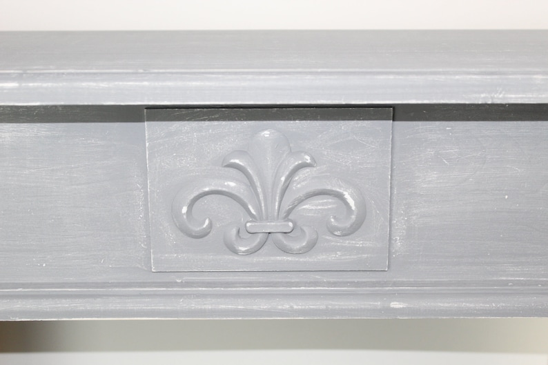 K&ouml;nnte beinhalten: Ein graues Holzregal mit einem dekorativen Fleur-de-lis-Design auf der Vorderseite. Das Regal hat ein schlichtes, elegantes Design.