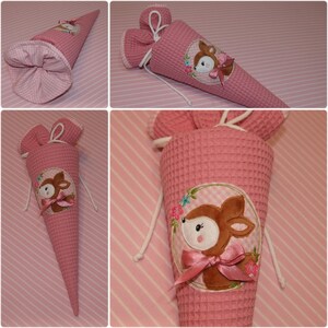 May include: A pink waffle-textured fabric cone with a drawstring closure at the top. The cone features an embroidered applique of a brown deer head inside a circular frame, accented with small colorful flowers and a pink satin ribbon bow.