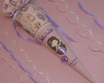 May include: A pink, cone-shaped school bag with a mermaid illustration and the name "Shirin". The bag is decorated with ribbons and a fabric top featuring mermaid and sea creature patterns. The text "Schulkind 2025" is also visible.