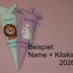 May include: Two cone-shaped fabric bags with drawstring closures. The left bag is light green with a lion graphic and the text Louis Kitakind 2026. The right bag is light purple with a unicorn graphic and the text Sophie Kitakind 2026.