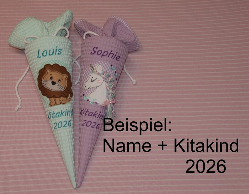 May include: Two cone-shaped fabric bags with drawstring closures. The left bag is light green with a lion graphic and the text Louis Kitakind 2026. The right bag is light purple with a unicorn graphic and the text Sophie Kitakind 2026.