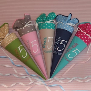 Könnte beinhalten: Fünf bunte, kegelförmige Schultüten mit sternförmigen Stoffoberteilen. Jede Tüte hat eine andere Farbe: Grün, Rosa, Beige, Marineblau und Türkis. Jede Tüte hat eine große weiße Zahl 5 und einen gestickten Namen.