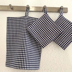 Könnte beinhalten: Ein Küchenset mit einem marineblauen und weißen Karomuster. Das Set umfasst ein rechteckiges Geschirrtuch und zwei quadratische Topflappen, die an einer Metallstange mit Haken hängen. Der Stoff hat ein klassisches Vichy-Design.