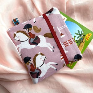 Könnte beinhalten: Ein rosa Buchhülle mit einem sich wiederholenden Muster eines Mädchens, das ein weißes Pferd reitet. Die Hülle hat ein rotes Gummiband und ein kleines Etikett. Ein grünes Buch ist teilweise sichtbar.