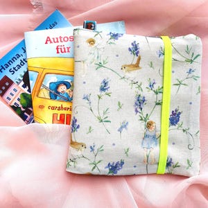 Könnte beinhalten: Ein Stoff-Buchcover mit blauem und grünem Blumenmuster, das drei Kinderbücher enthält. Das Cover hat ein leuchtend gelbes Gummiband. Die Bücher haben deutsche Titel.