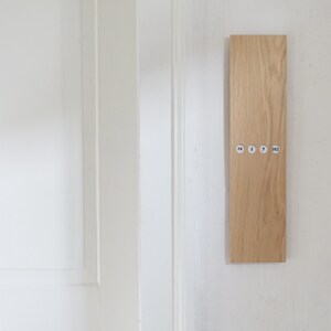 May include: A rectangular wooden wall calendar with the text "FR 2 7 DEZ" printed on small circular stickers. The calendar is made of light-colored wood and is mounted on a white wall next to a white door.