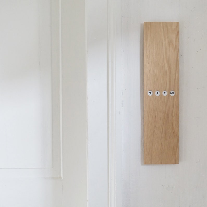 May include: A rectangular wooden wall calendar with the text "FR 2 7 DEZ" printed on small circular stickers. The calendar is made of light-colored wood and is mounted on a white wall next to a white door.
