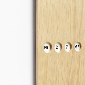 May include: Close-up of a wooden calendar with the date "FR 2 7 DEZ" displayed in white circular inserts. The wood has a light brown color and visible grain. The background is white.