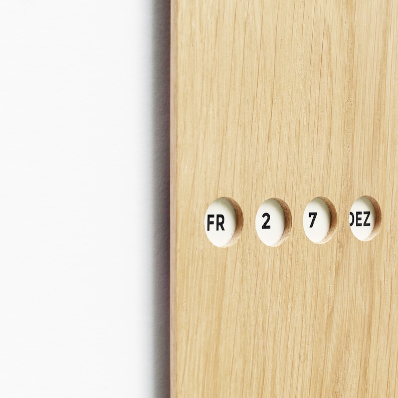 May include: Close-up of a wooden calendar with the date "FR 2 7 DEZ" displayed in white circular inserts. The wood has a light brown color and visible grain. The background is white.