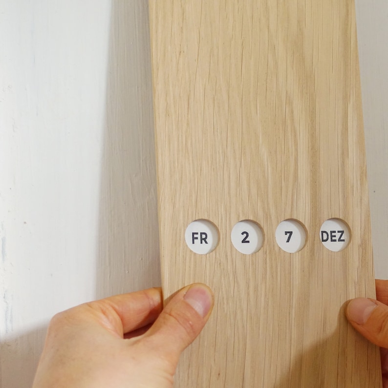 May include: A light-colored wooden calendar with the day and date displayed in white circles. The text on the circles reads "FR 2 7 DEZ". The calendar is held by two hands against a white wall.