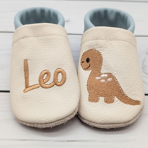 Könnte beinhalten: Ein Paar cremefarbene Leder-Babyschuhe mit hellblauem Rand. Der linke Schuh zeigt den gestickten Namen Leo in braunem Garn, während der rechte Schuh einen gestickten braunen Dinosaurier mit weißen Flecken auf dem Rücken aufweist.