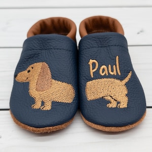 Può includere: Un paio di scarpine da bambino in pelle blu navy con finiture marroni. Ogni scarpa presenta un ricamo di bassotto color camoscio. Una scarpa è ricamata con il nome "Paul" in filo color camoscio. Le scarpe sono appoggiate su una superficie di legno bianco.