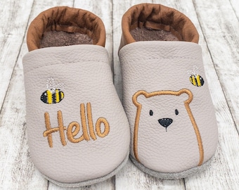 May include: A pair of gray baby shoes with brown trim. One shoe has the word "Hello" embroidered on it, and the other has a bear face embroidered on it. Both shoes have a bee embroidered on them.