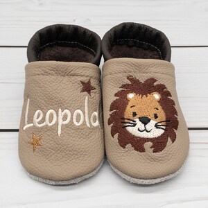Op de afbeelding: Een paar beige leren babyschoentjes met geborduurde sterren en de naam "Leopold" op de ene schoen en een leeuwenkop op de andere.