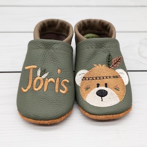 Può includere: Un paio di scarpine da bambino verde oliva con suole marroni e interno marrone. Una scarpa ha il nome "Joris" ricamato in oro, e l'altra presenta una faccia di orso con una piuma e una fascia.