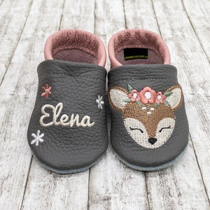 Könnte beinhalten: Ein Paar graue Lederbabyschuhe. Ein Schuh zeigt den gestickten Namen "Elena" mit floralen Akzenten. Der andere Schuh hat ein gesticktes Rehkitzgesicht mit einem Blumenkranz. Das Innenfutter ist rosa.