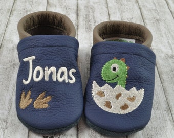 May include: A pair of blue leather baby shoes with a dinosaur design on one shoe and the name "Jonas" embroidered on the other shoe.