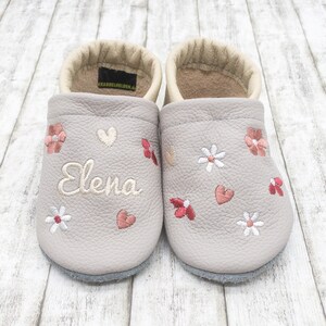 Op de afbeelding: Een paar grijze leren babyschoentjes met geborduurde bloemen, hartjes en de naam "Elena".