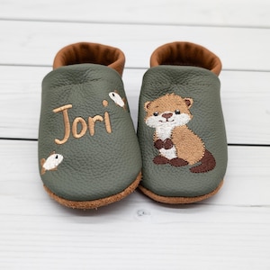 Könnte beinhalten: Ein Paar olivgrüne Leder-Babyschuhe mit braunem Rand. Der linke Schuh ist mit dem Namen Jori in braunem Garn und zwei kleinen weißen Fischen bestickt. Der rechte Schuh zeigt eine gestickte braun-weiße Otterfigur als Motiv.