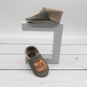 May include: Two baby moccasins. One is olive green and tan with a lion face embroidered on the toe. The other is olive green and tan with a lion face embroidered on the toe. The shoes are made of leather.