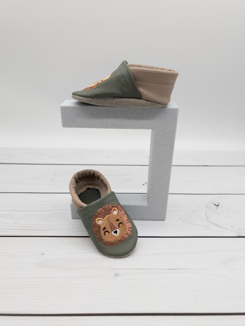 May include: Two baby moccasins. One is olive green and tan with a lion face embroidered on the toe. The other is olive green and tan with a lion face embroidered on the toe. The shoes are made of leather.