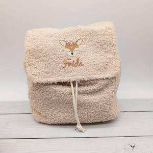 Op de afbeelding: Een beige, pluizige rugzak met een trekkoordsluiting. De voorkant heeft een geborduurd ontwerp van een hert met een bloemenkroon en de naam "Frida" in goudkleurig garen. De rugzak is gemaakt van een zacht, getextureerd materiaal.