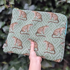 Könnte beinhalten: Eine grüne Reißverschlusstasche mit einem sich wiederholenden Muster aus braunen Leoparden auf grünem Hintergrund. Die Tasche hat einen blauen Reißverschluss und das Wort "HILO" ist auf der Vorderseite aufgedruckt.